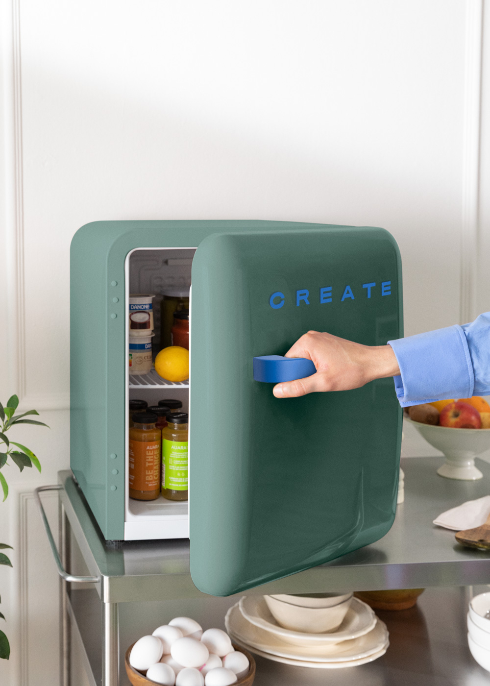 Un mini-refrigerador verde azulado abierto, revela alimentos. Una mano abre la puerta. Huevos y fruta cerca.