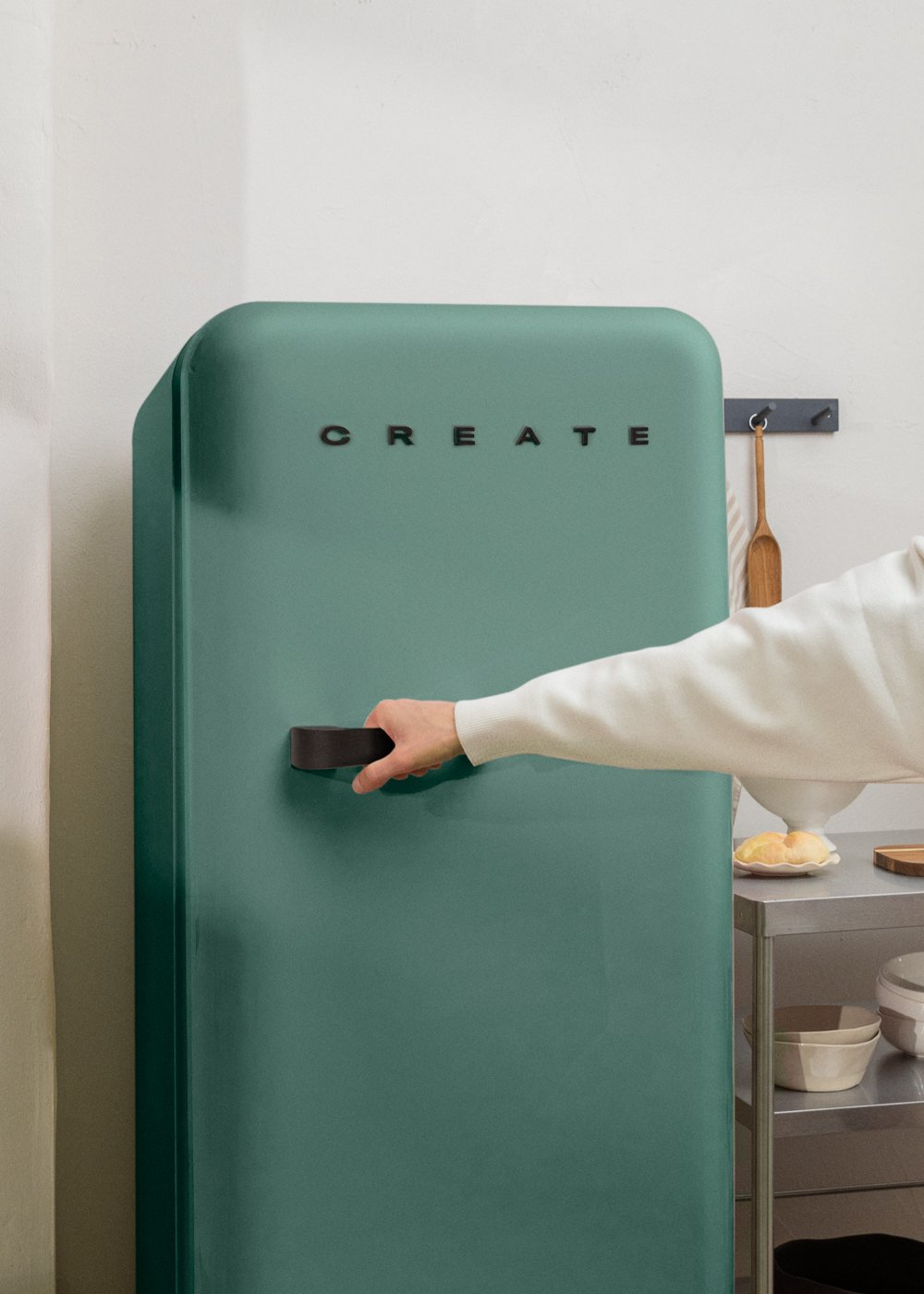 Una mano abre un refrigerador verde azulado con la palabra CREATE en la puerta. Se ve una cuchara y un plato.