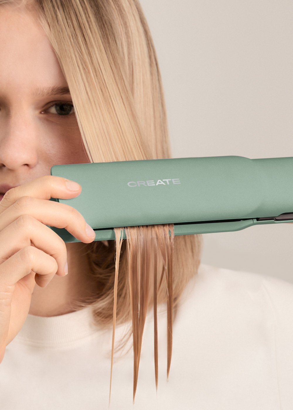 Una persona usa una plancha verde, mechones de cabello entre las placas. Fondo claro.