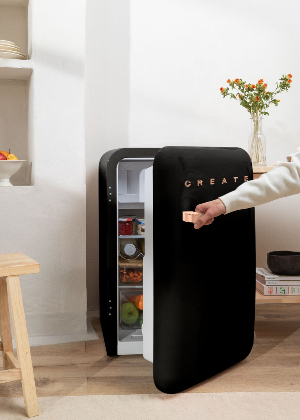 Un mini-refrigerador negro abierto, muestra alimentos. Una mano abre la puerta. Un taburete y flores cerca.