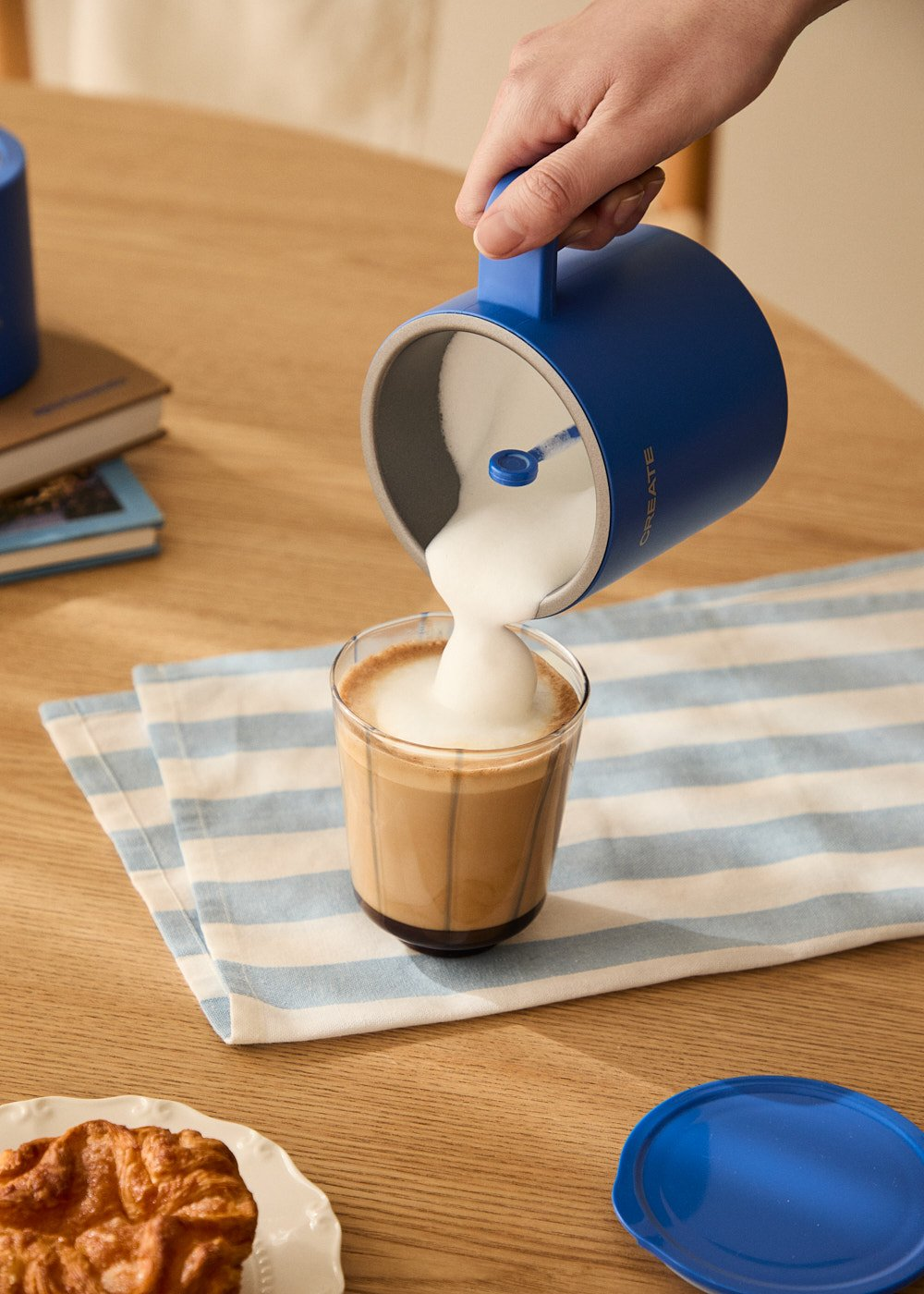 Una persona vertiendo leche espumada en un vaso de café con un espumador azul.