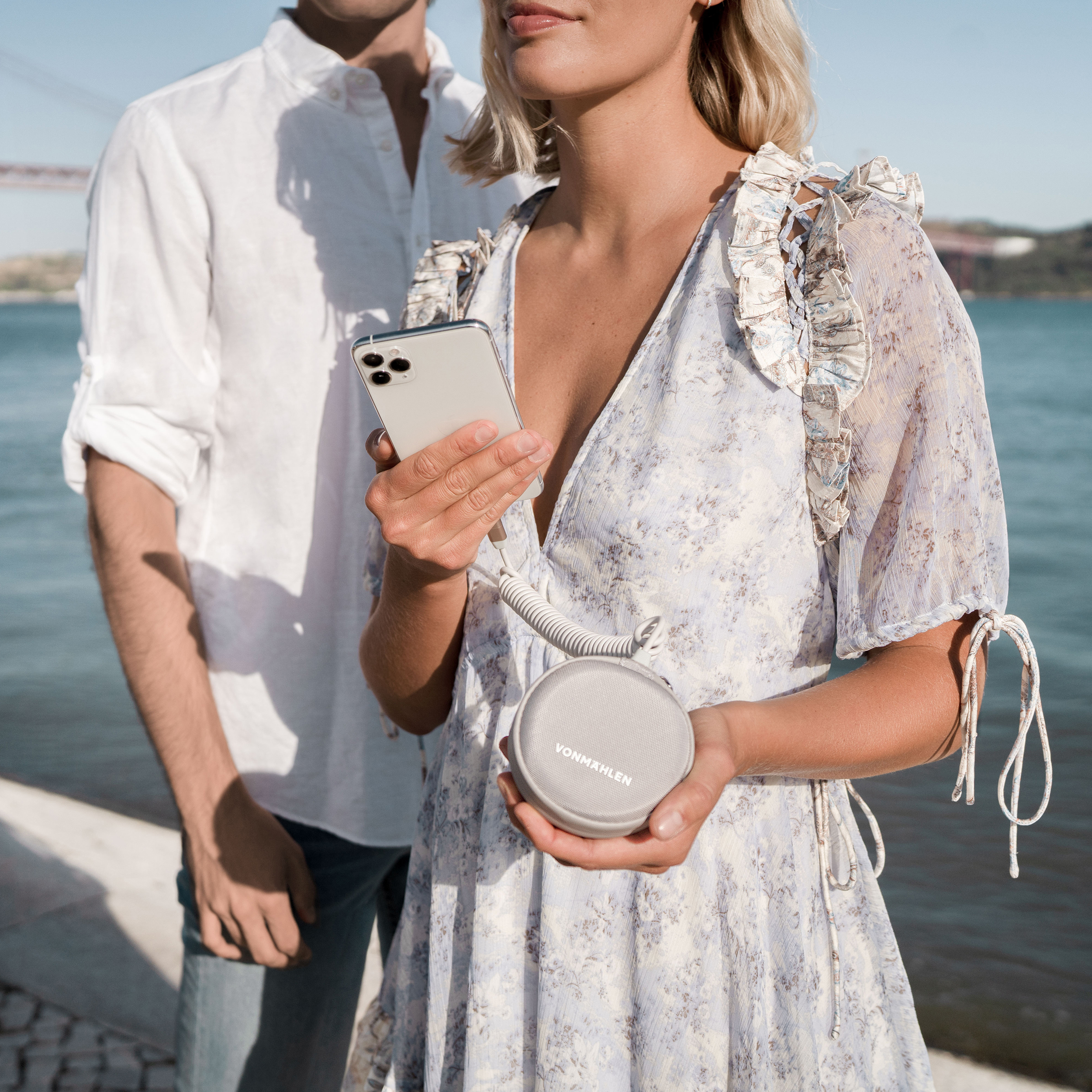 Ein Paar mit Telefon und Lautsprecher. Sie stehen am Wasser, mit einer Brücke im Hintergrund.