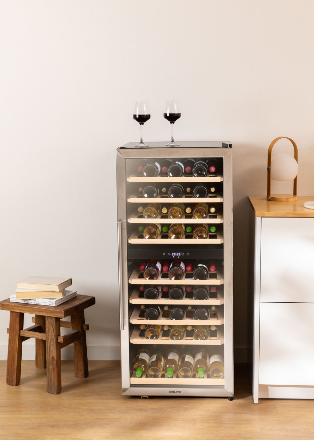 Enfriador de vino con botellas de vino. Dos copas de vino encima. Taburete de madera y armario blanco a los lados.