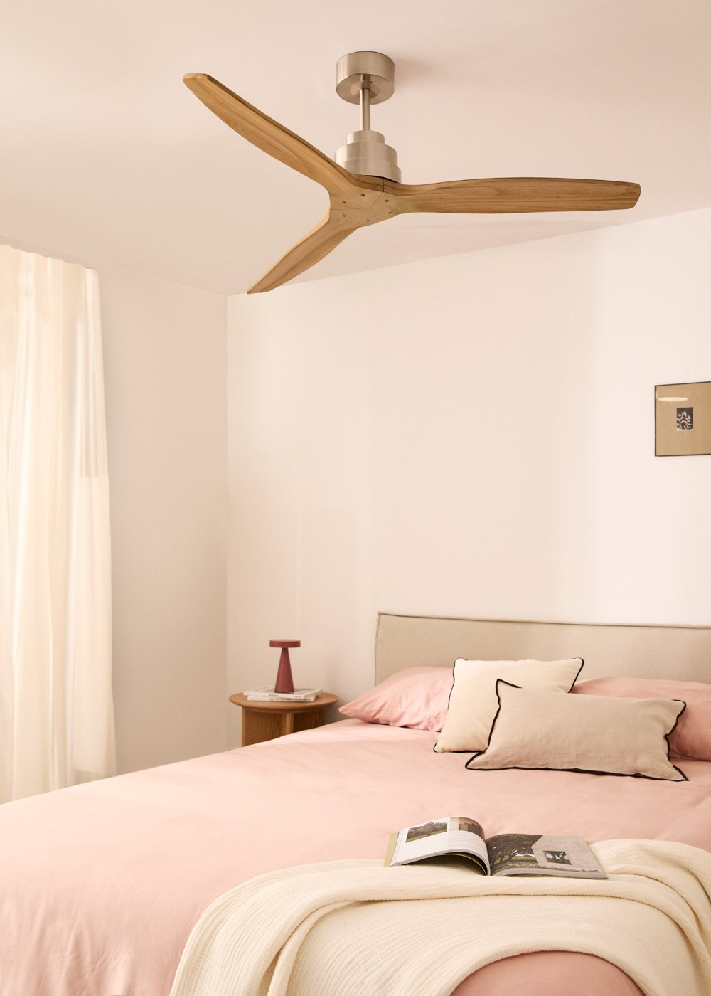 Dormitorio con ventilador de techo de madera, cama rosa, paredes blancas y ventana con cortina.