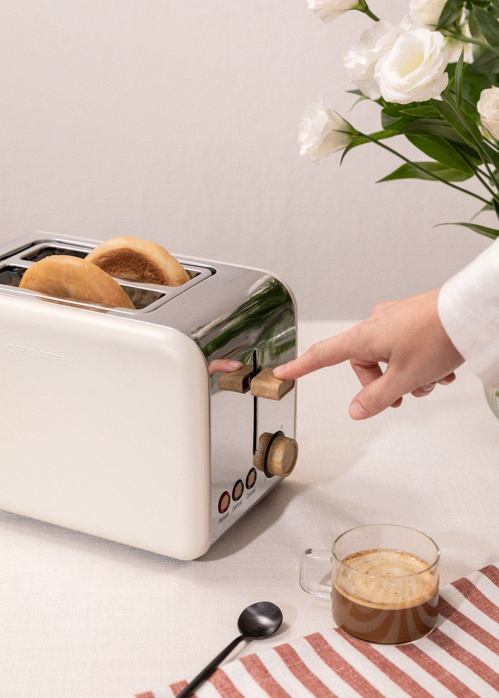 Una tostadora blanca con dos rebanadas de pan tostado. Una mano empuja la palanca hacia abajo. Hay una taza de café cerca.