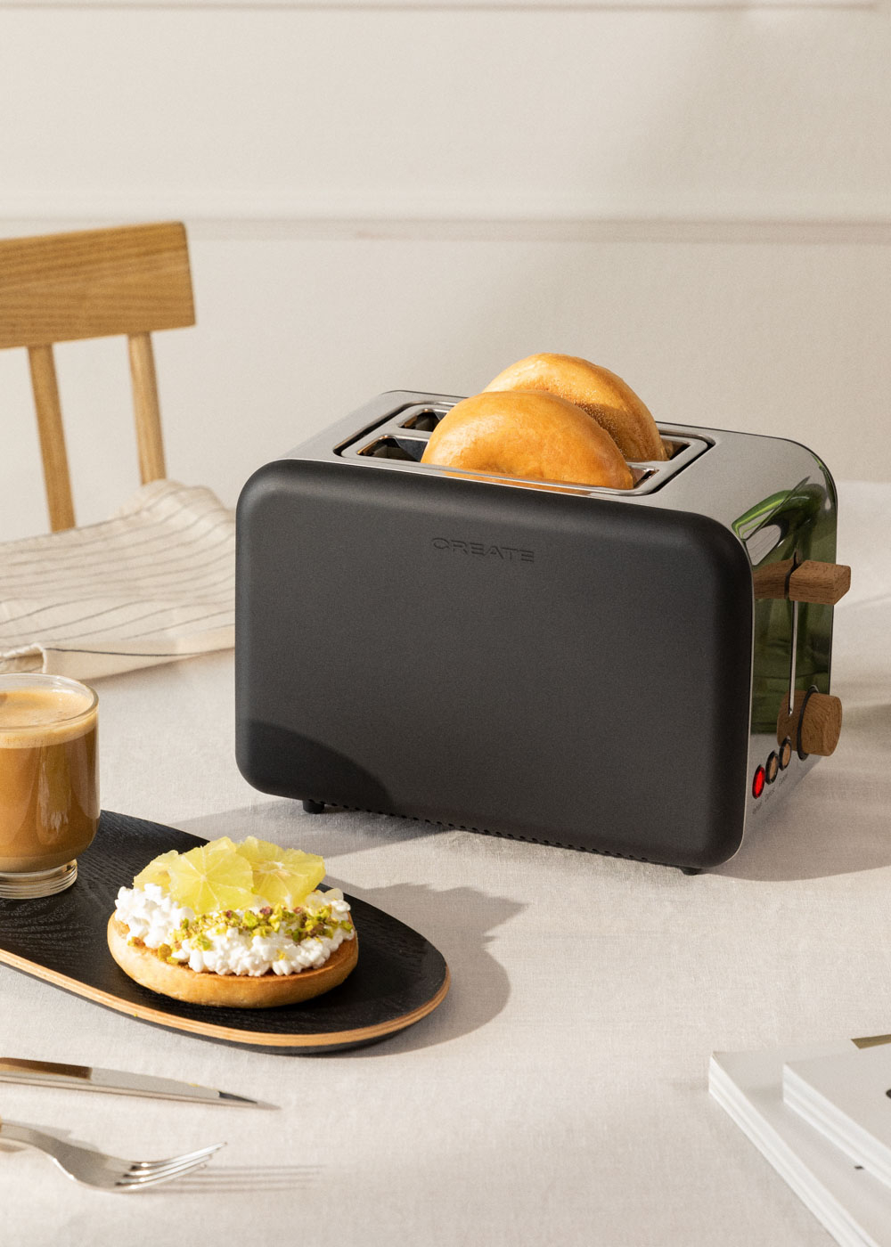 Una tostadora negra con pan dentro. Se muestra una mesa con un plato de comida y una taza de café.