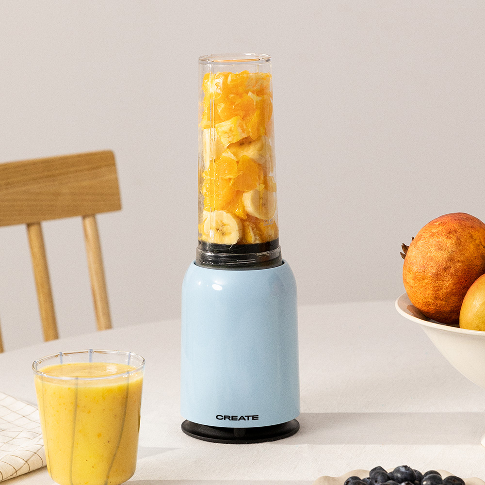 Ein blauer Mixer, gefüllt mit Orangenscheiben und Bananenscheiben. Daneben steht ein Glas Smoothie.