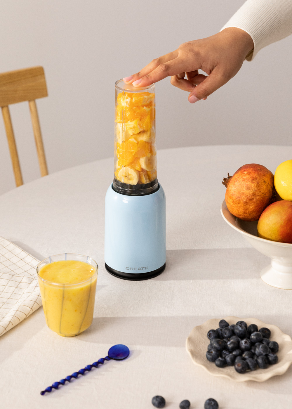 Eine Person drückt einen Knopf an einem blauen Mixer. Zutaten sind Orangen und Bananen.