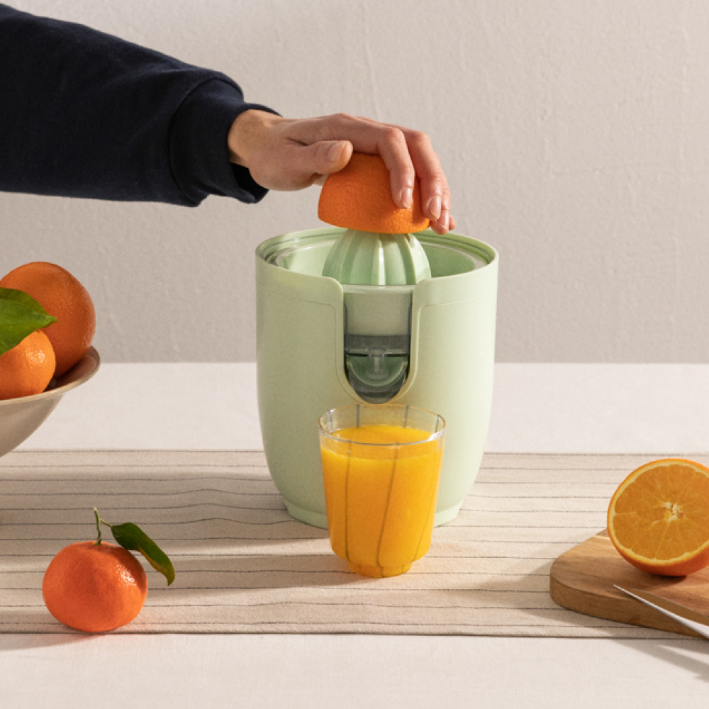 Persona exprimiendo media naranja en exprimidor. Hay un vaso de zumo y naranjas.