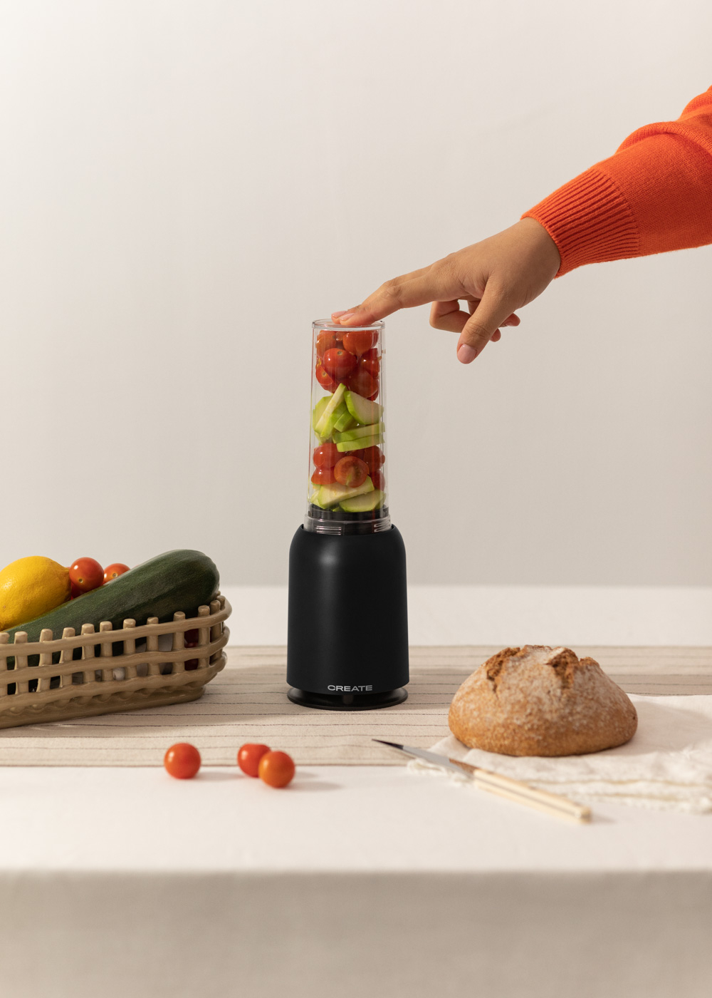 Una persona pulsa un botón en una licuadora con verduras dentro. Pan y tomates en la mesa.