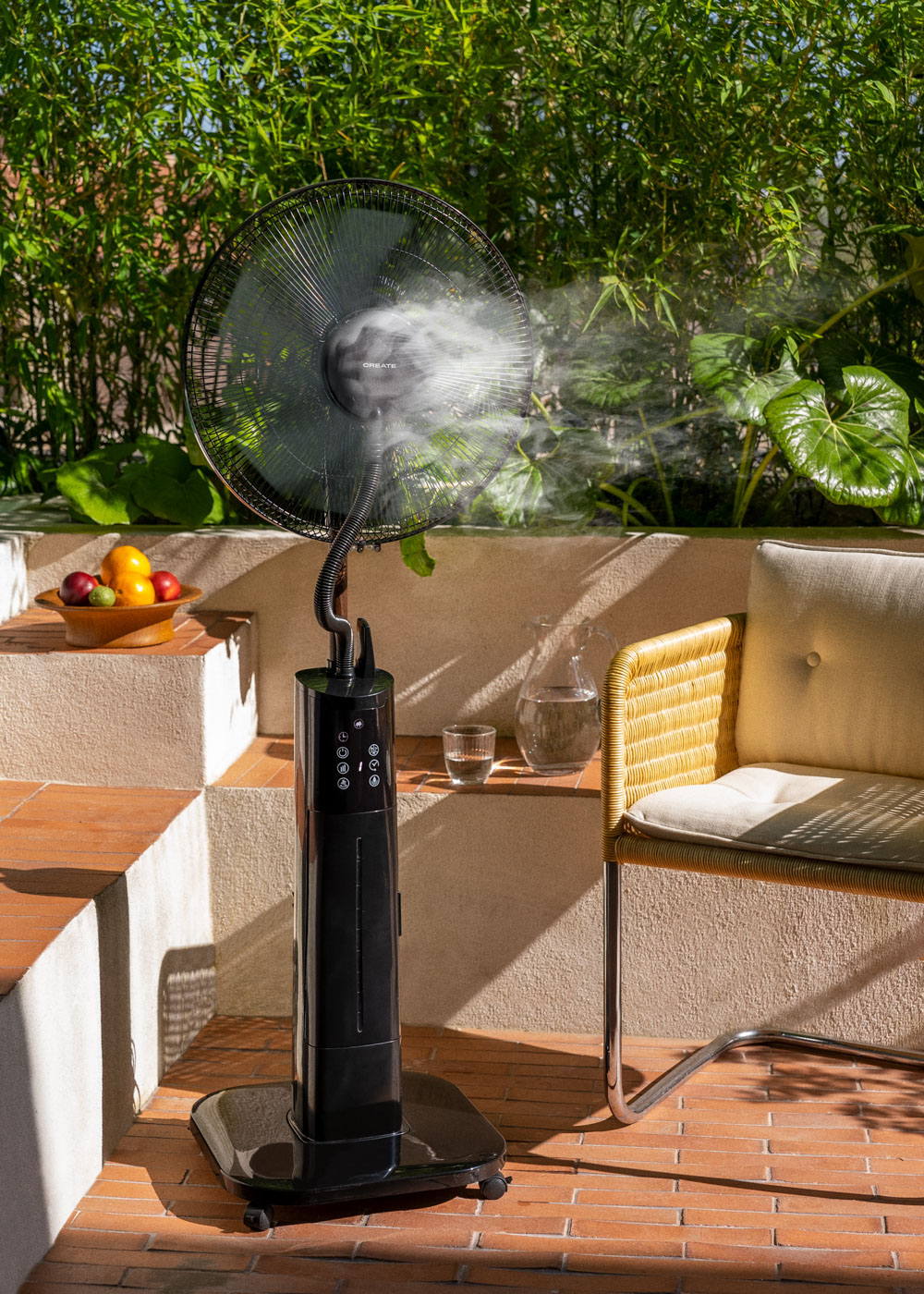Ventilador negro con niebla. Frutero en escalones. Entorno al aire libre, plantas al fondo.