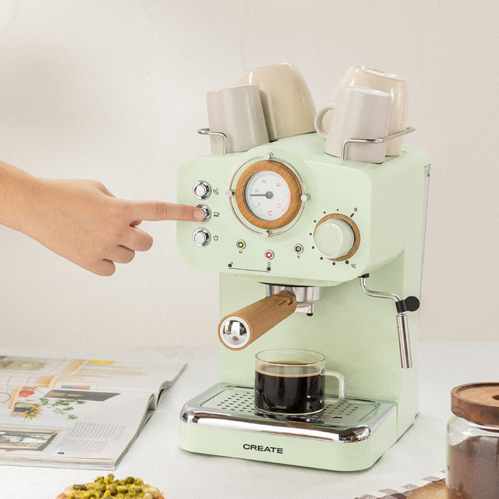 Máquina de espresso verde menta con una mano pulsando un botón. Tiene tazas encima y un vaso de café.