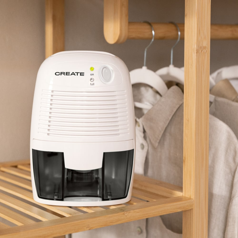 Deshumidificador CREATE blanco. Estante de madera con ropa colgando. El deshumidificador tiene una luz 'ON'.