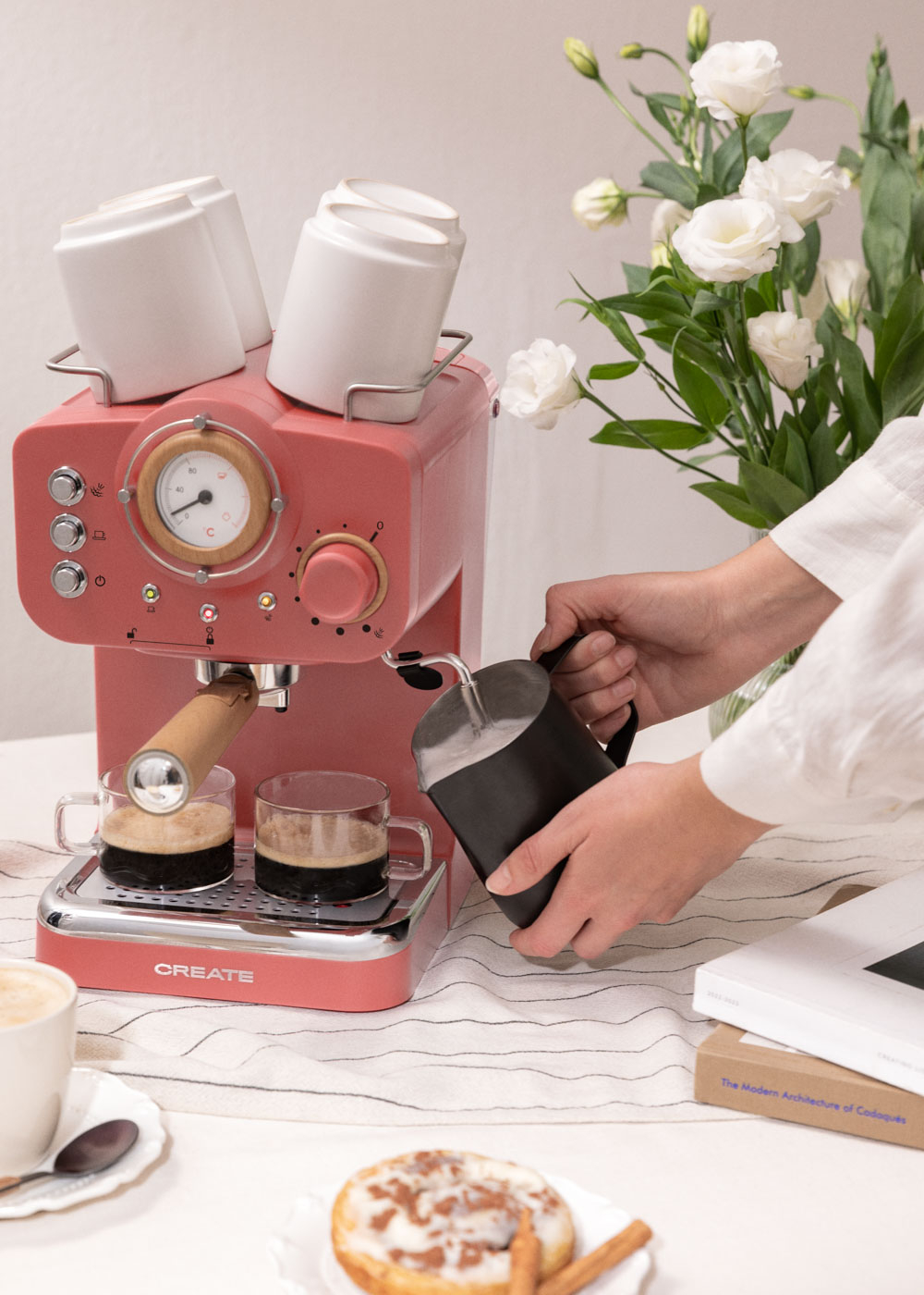 Una persona vertiendo agua en una máquina de espresso rosa con tazas encima. Tazas de café y una dona están en la mesa.