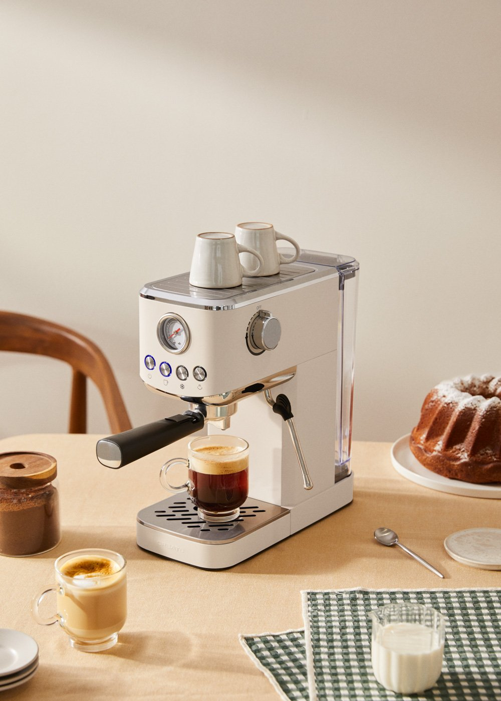 Una máquina de espresso blanca y plateada con tazas encima. Hay café y un pastel cerca.