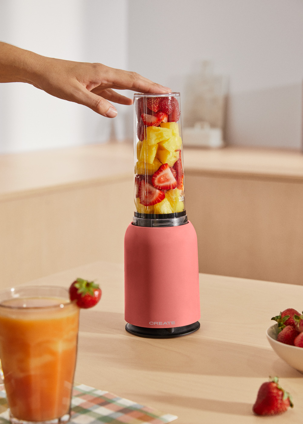 Una licuadora rosa con fruta dentro, una mano se acerca. Un vaso de jugo a la izquierda.