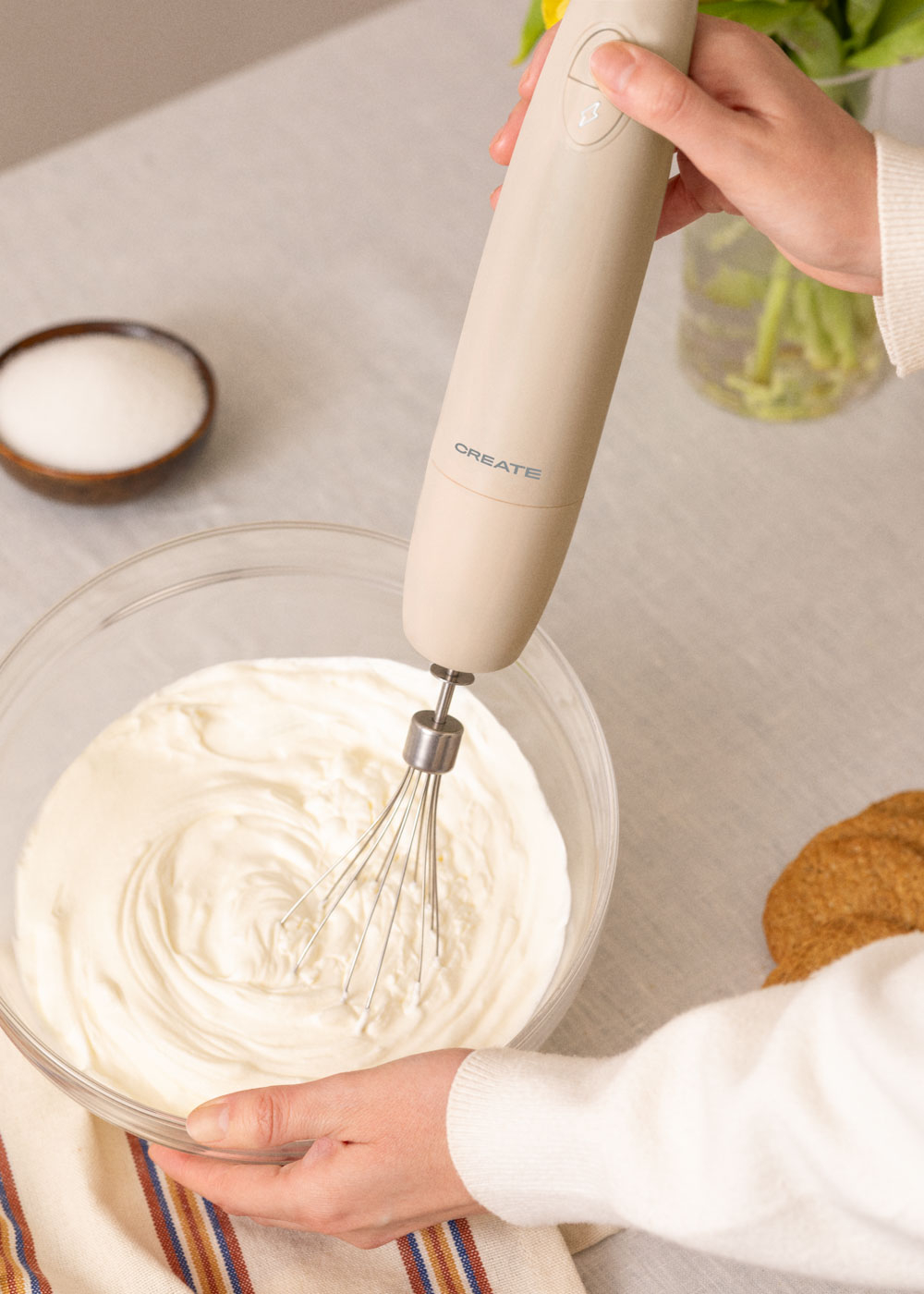 Una mano sosteniendo una batidora de mano beige sobre un cuenco de vidrio con crema batida, azúcar y plato.