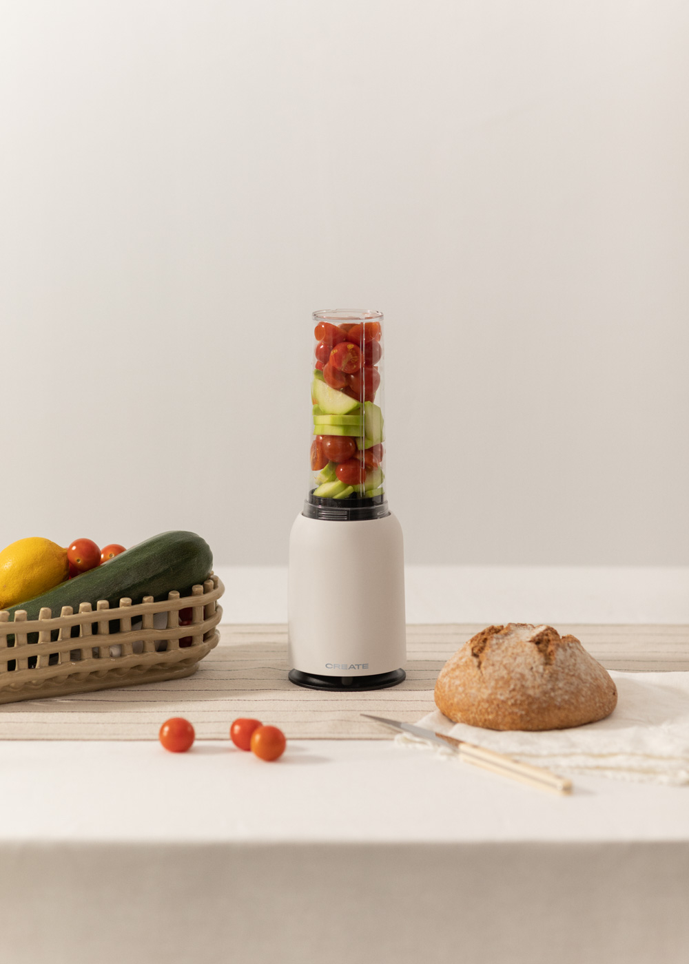 Ein Mixer mit Tomaten und Gurken darin. Ein Korb mit Zitronen und Brot steht auf dem Tisch.