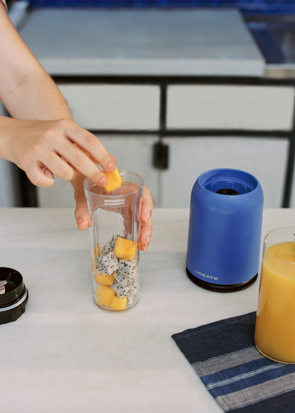 Persona agregando fruta a un vaso alto con otras frutas y una licuadora.