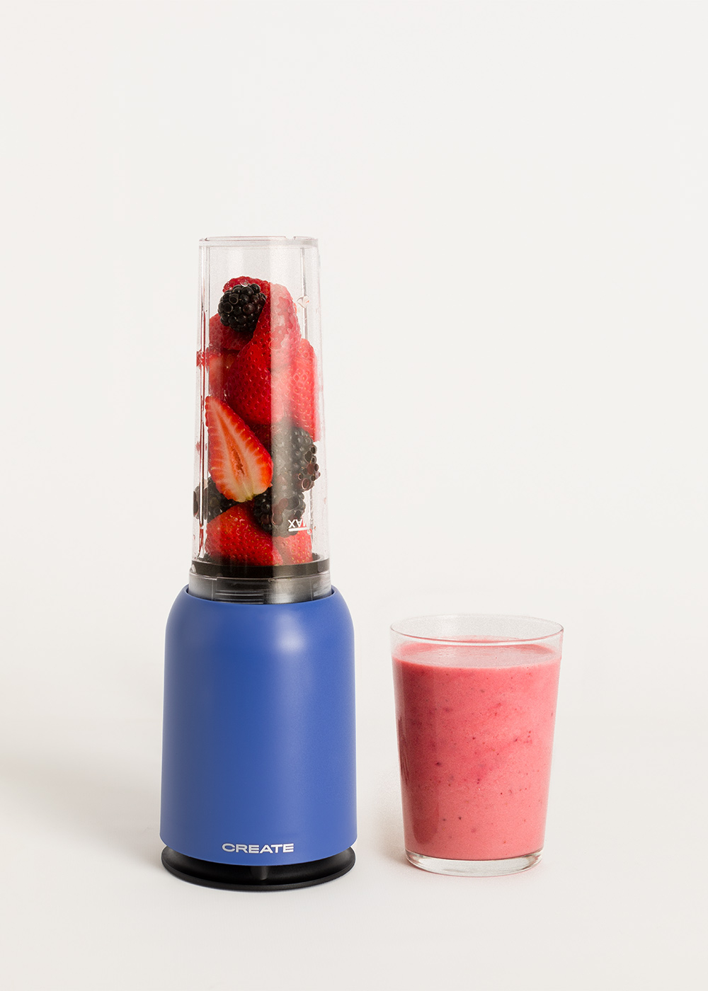 Licuadora azul llena de bayas, junto a un vaso de batido rosa.