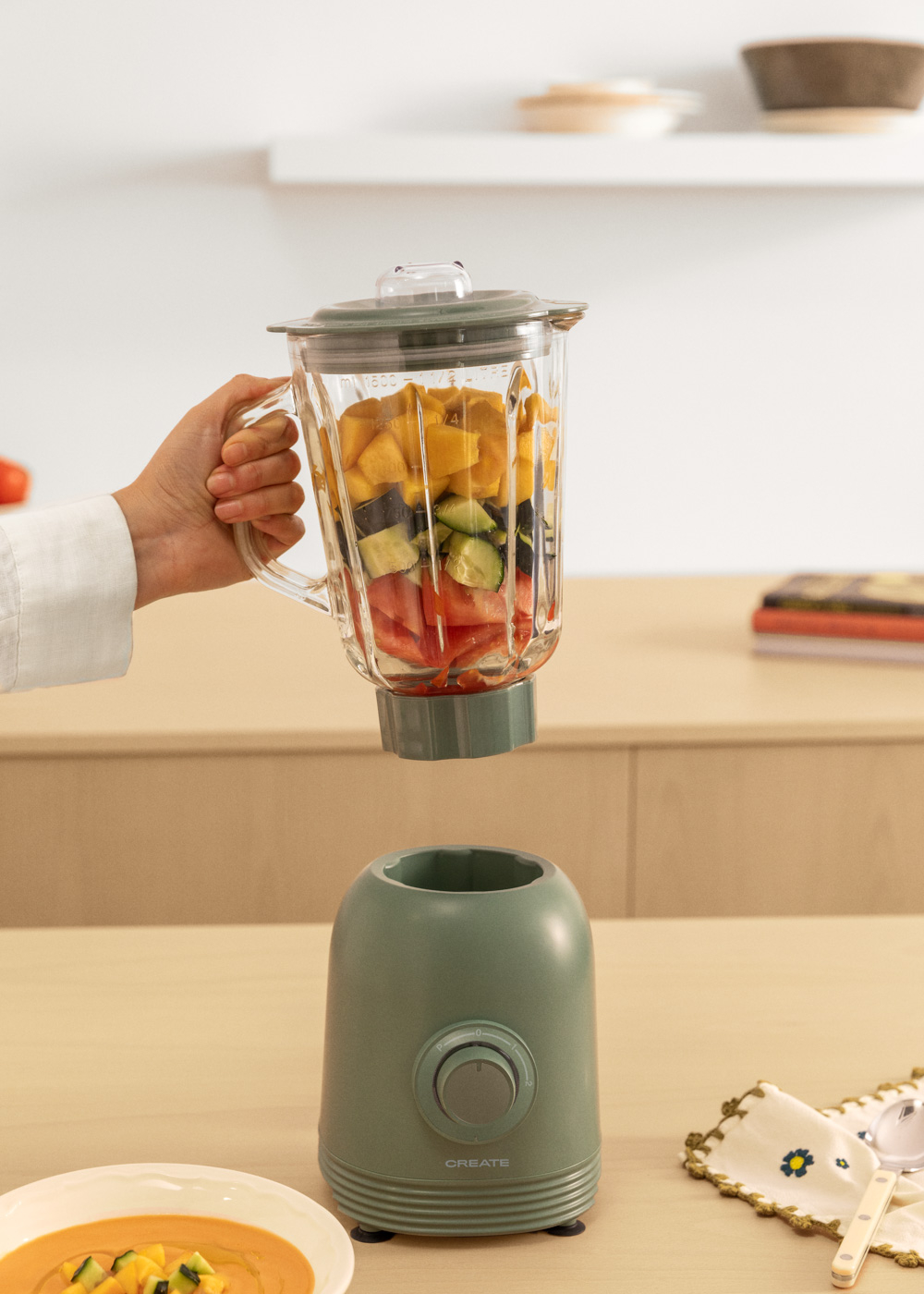 Person hält Mixer mit geschnittenem Gemüse. Tisch mit Gegenständen. Weißer Raum.