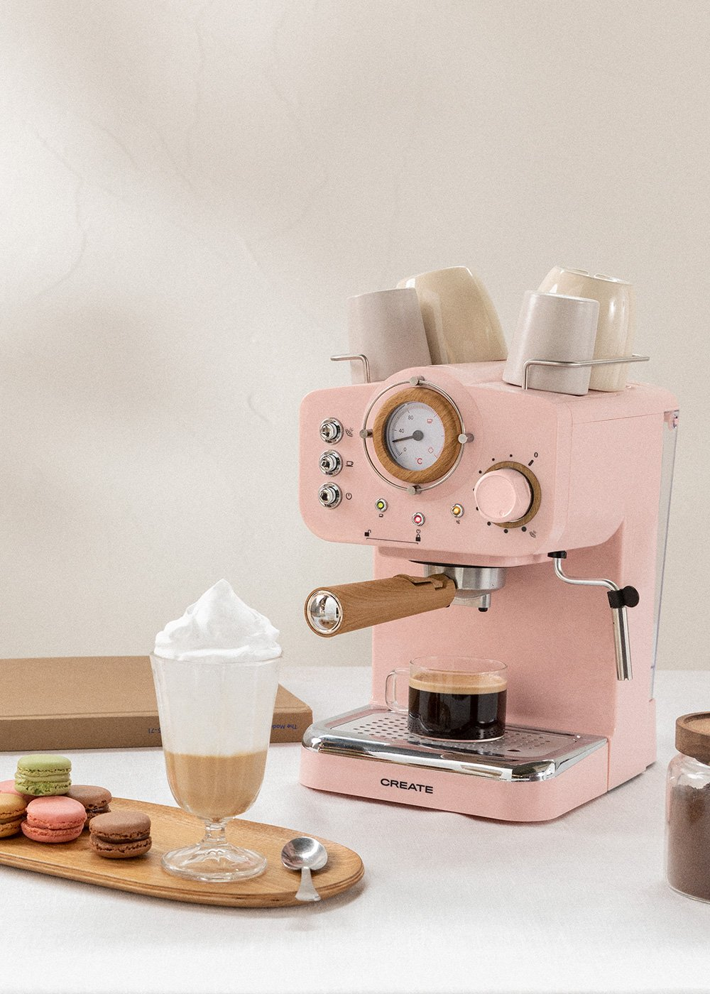 Máquina de espresso rosa, bandeja de madera con café, vaso de latte, macarons en mesa.