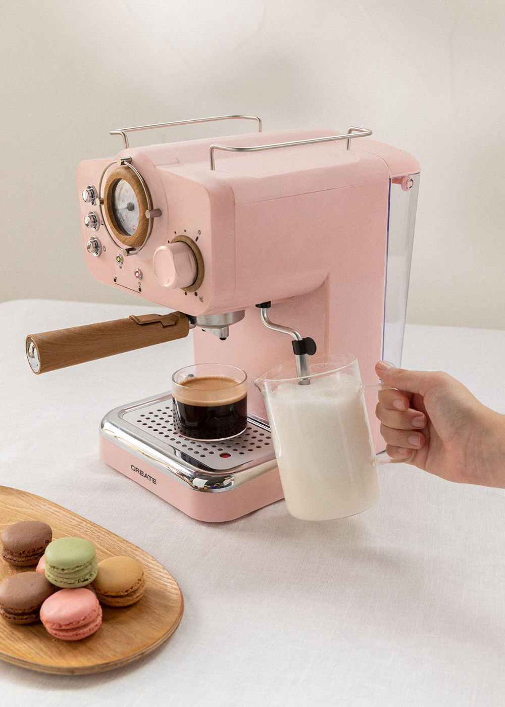 Máquina de espresso rosa con un vaso de café y macarons. Una mano vierte leche.