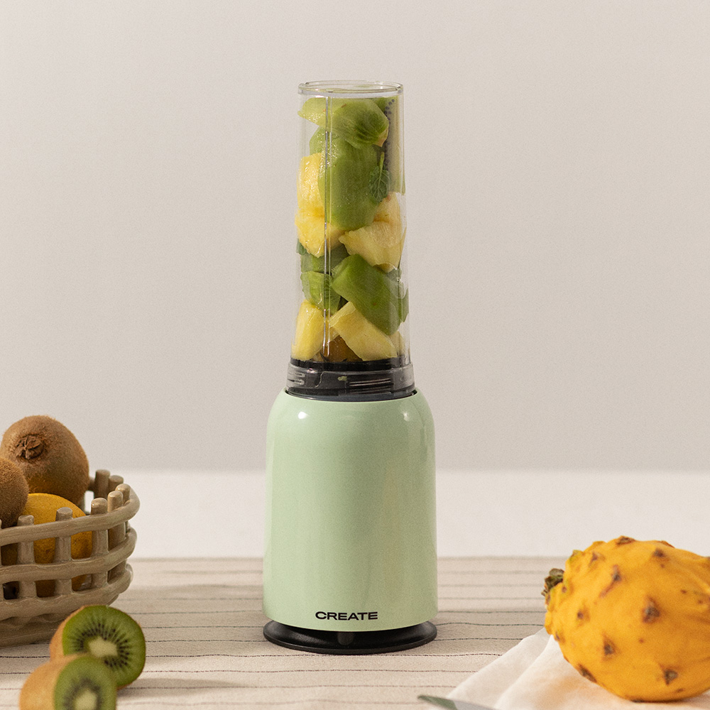 Una licuadora verde llena de frutas picadas, con kiwis y una fruta del dragón al lado.