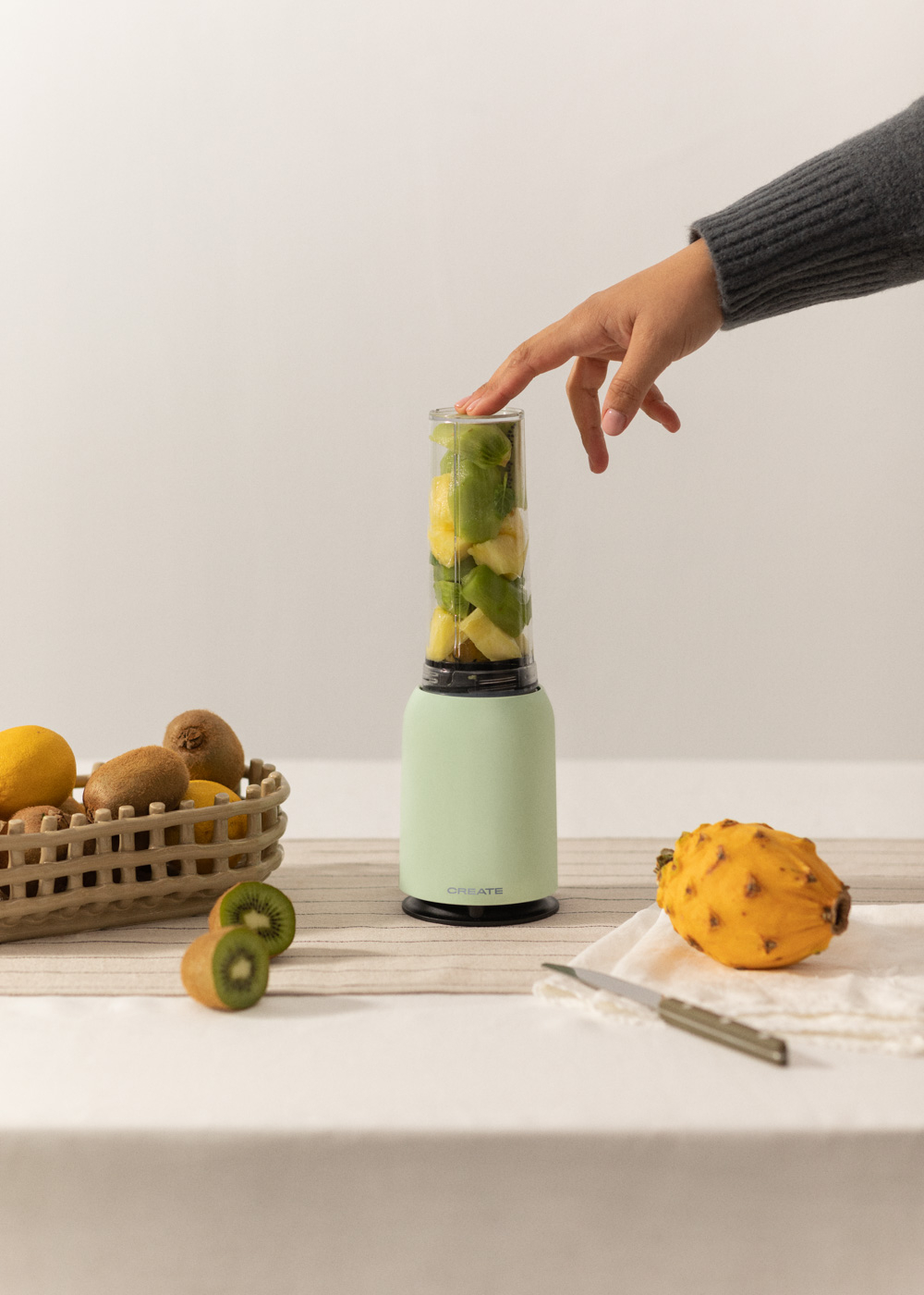 Mano presionando la parte superior de una licuadora llena de fruta. Kiwi y limón delante.