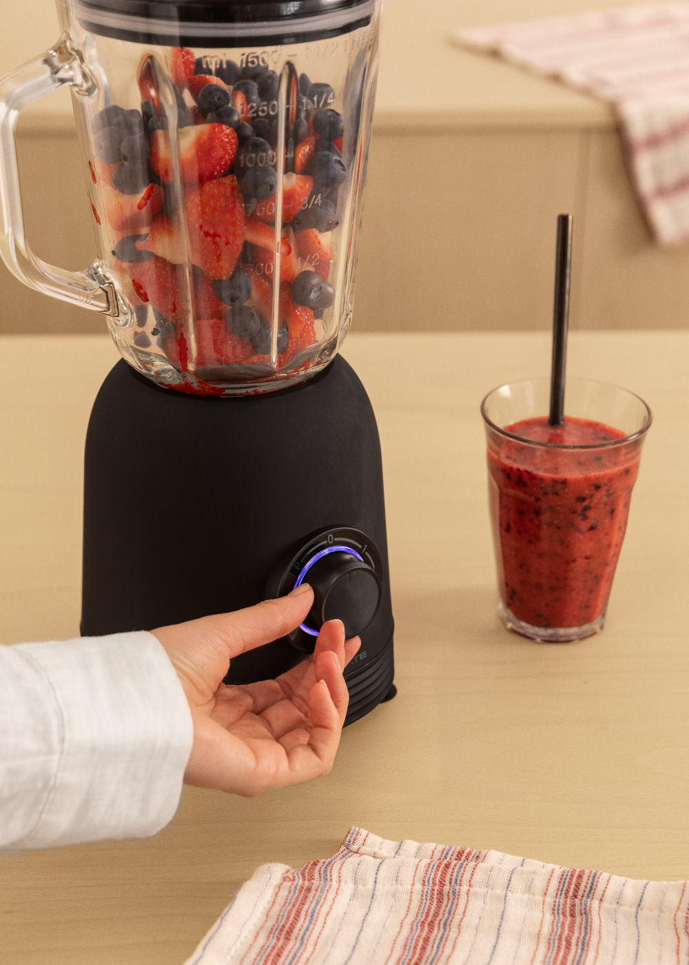 Eine Person dreht den Drehregler an einem schwarzen Mixer, der mit Beeren und Erdbeeren gefüllt ist.