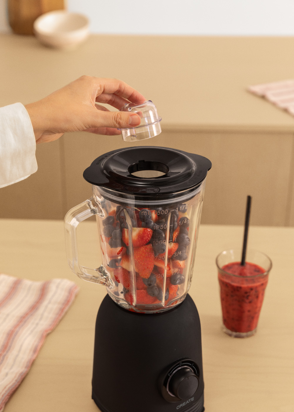 Mixer mit Früchten, Hand legt Deckel auf. Smoothie-Glas daneben.
