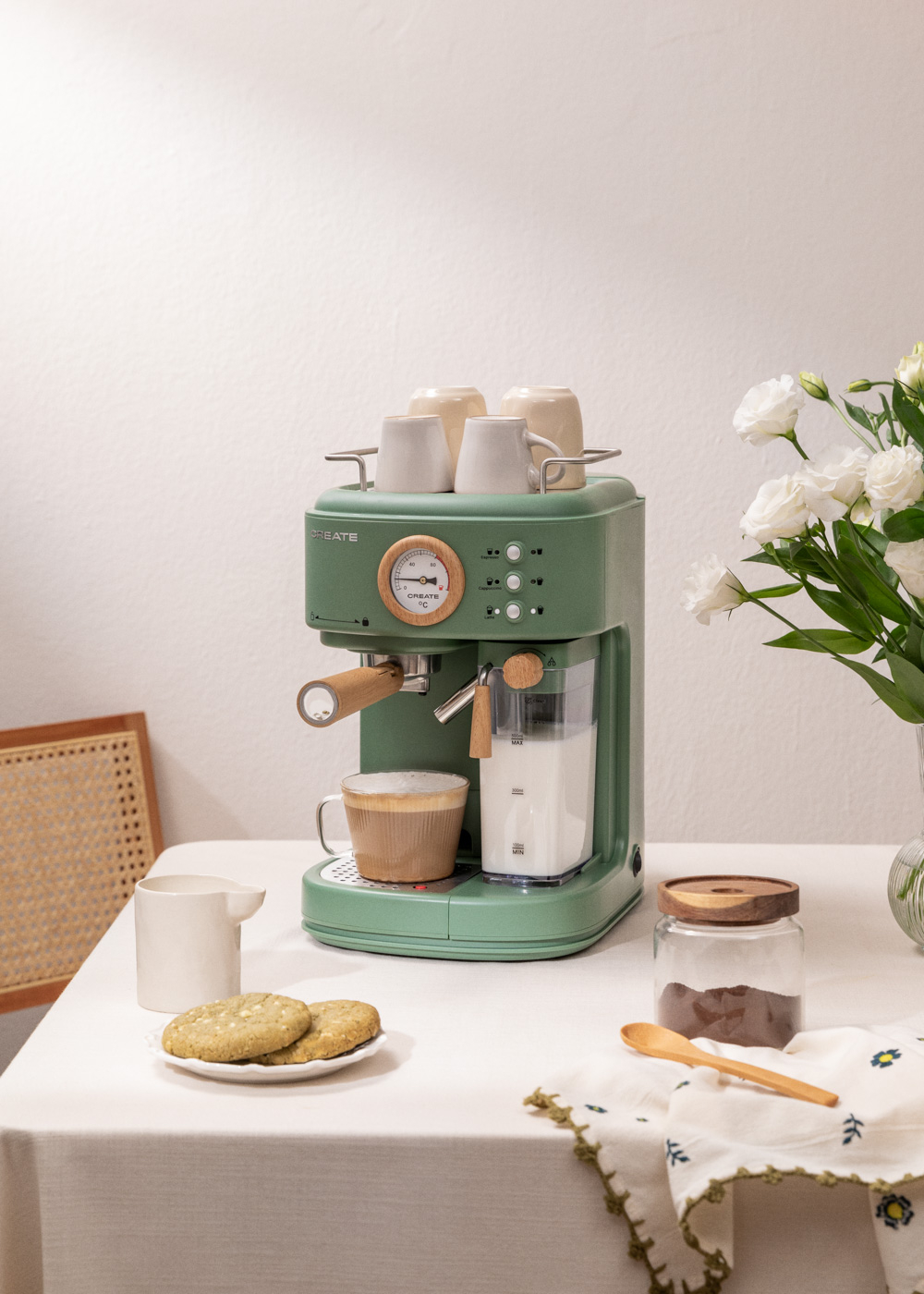 Máquina de espresso verde, jarra de leche, taza de café, galletas, flores y mesa.