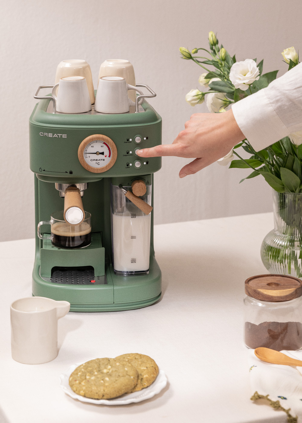 Una persona presionando un botón en una cafetera verde con tazas y galletas.