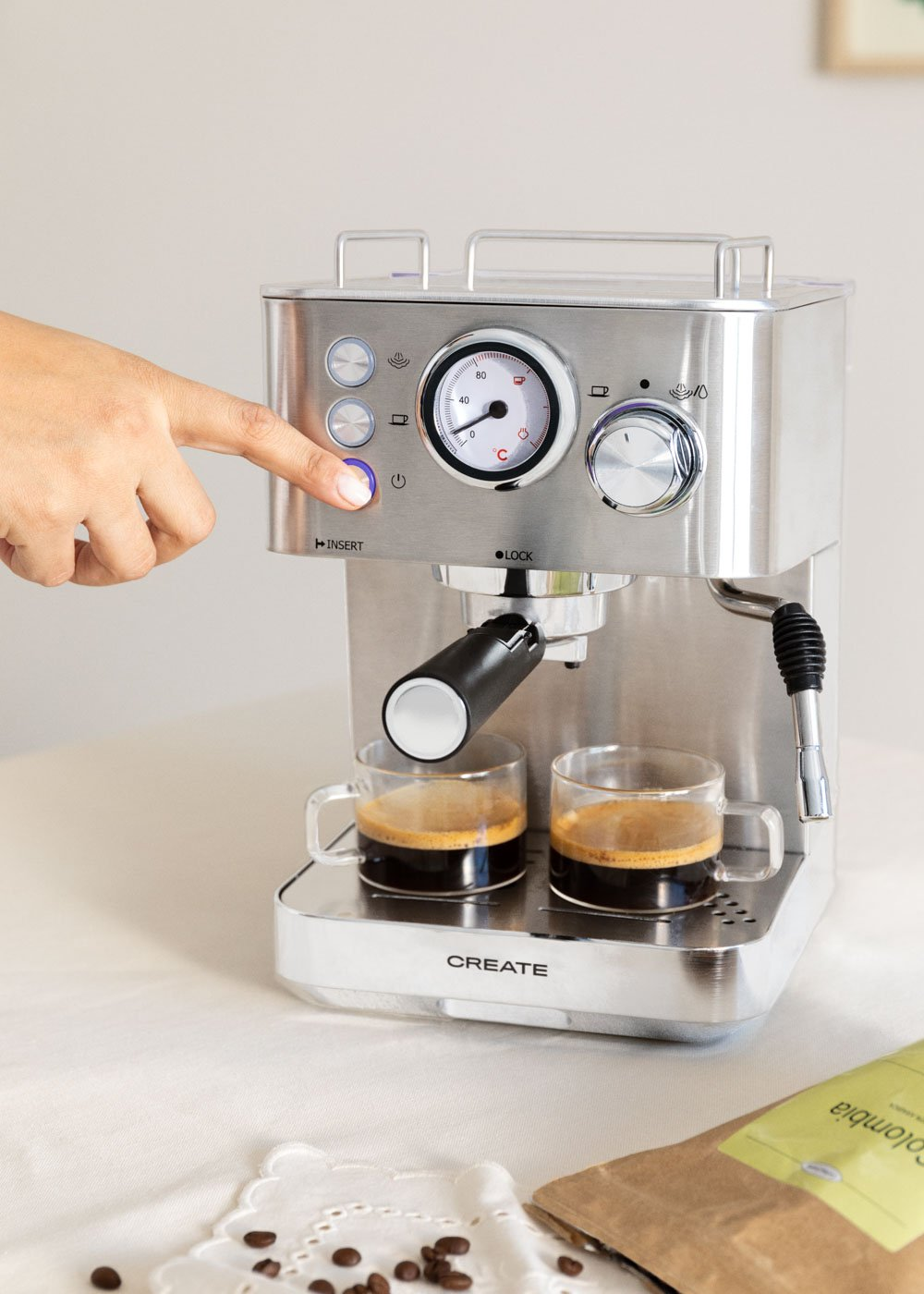Una persona presiona un botón en una máquina de espresso plateada. Dos tazas de café en la bandeja.