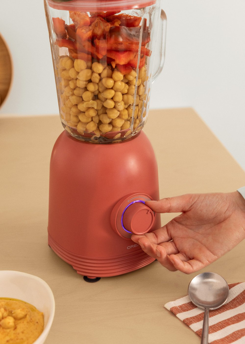 Hand dreht Regler an pinkem Mixer mit Kichererbsen und roten Paprika im Inneren.