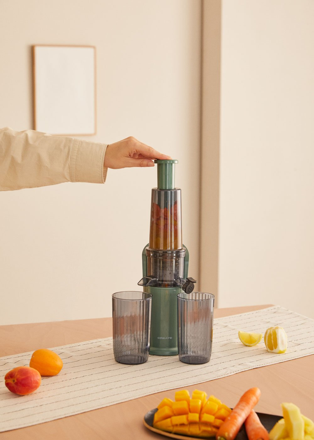 Exprimidor CREATE verde con dos vasos. Mano operándolo. Zanahorias, mango y limón en la mesa.