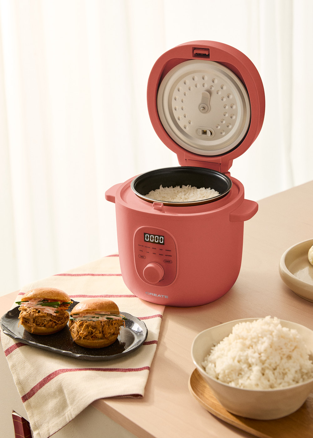 Una arrocera rosa con la tapa abierta, mostrando arroz. Se ven dos deslizadores en un plato y arroz en un cuenco.
