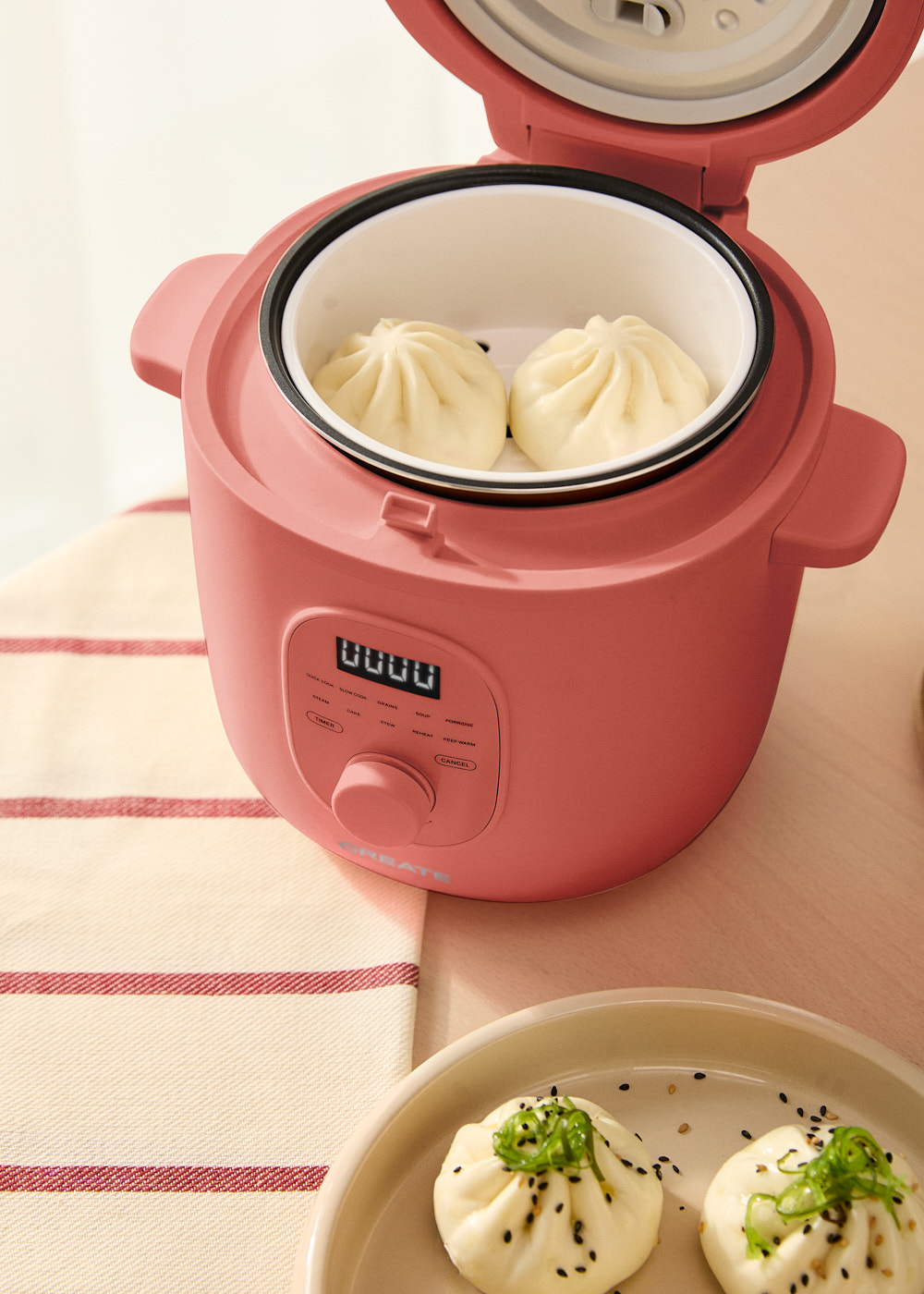 Cocina eléctrica rosa rojiza con dos bollos al vapor dentro. Plato con bollos delante.