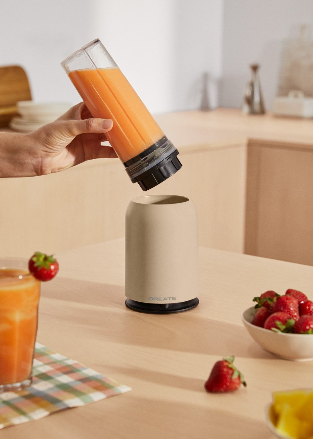 Hand gießt Orangensaft in eine beige Blenderbasis. Erdbeeren und ein Saftglas stehen auf einem Holztisch.