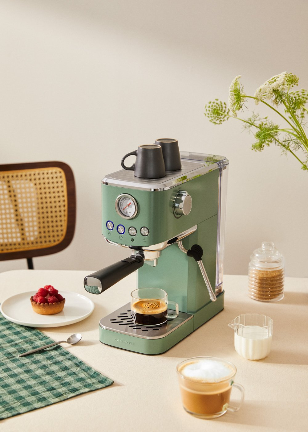 Una máquina de espresso verde con tazas, una tarta y azúcar, en una mesa blanca.