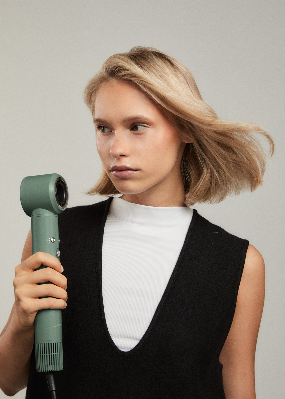 Mujer sosteniendo un secador de pelo verde. Cabello frente al secador. Fondo gris claro.