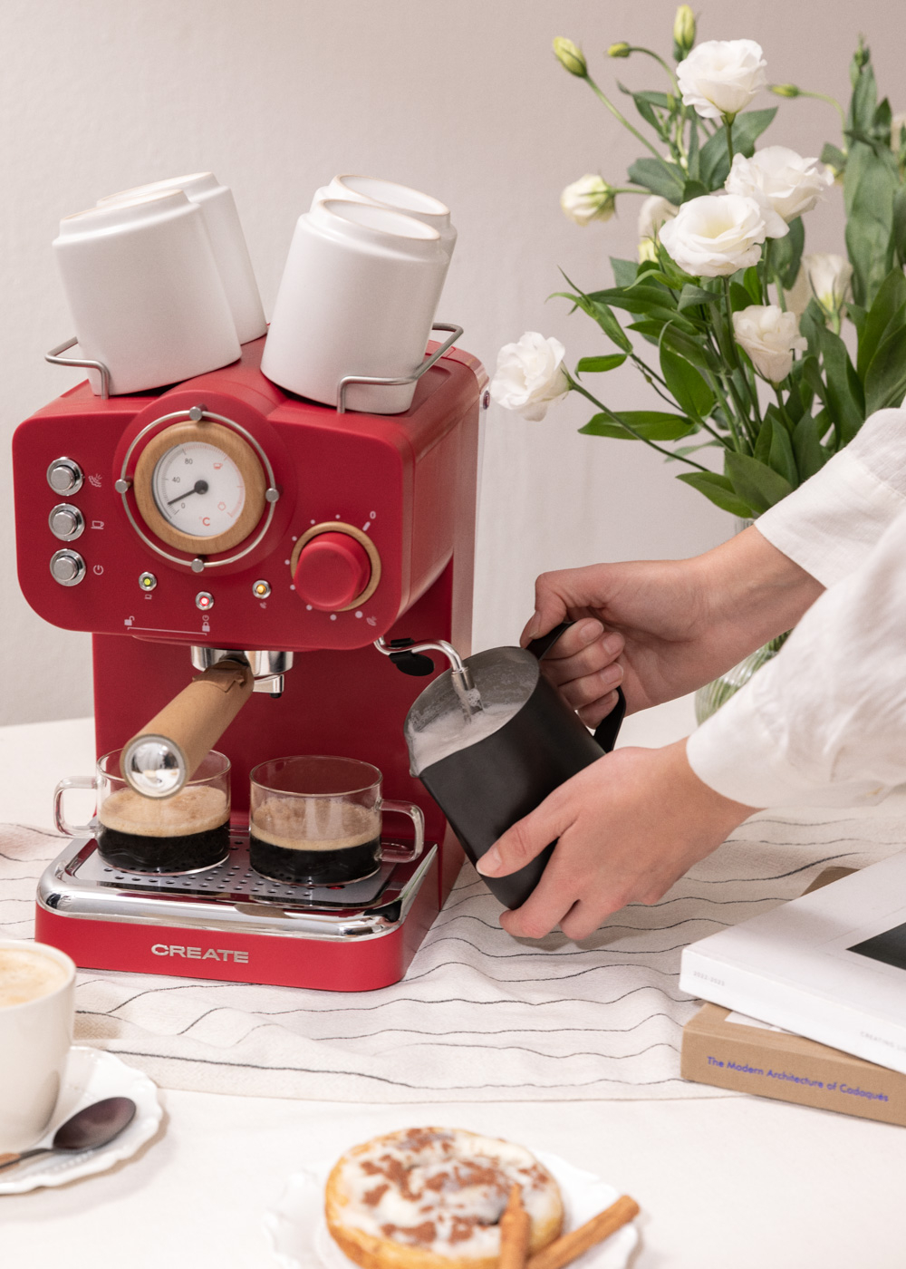 Máquina de espresso roja sirviendo café. Tazas blancas y flores en la mesa, una persona vierte.