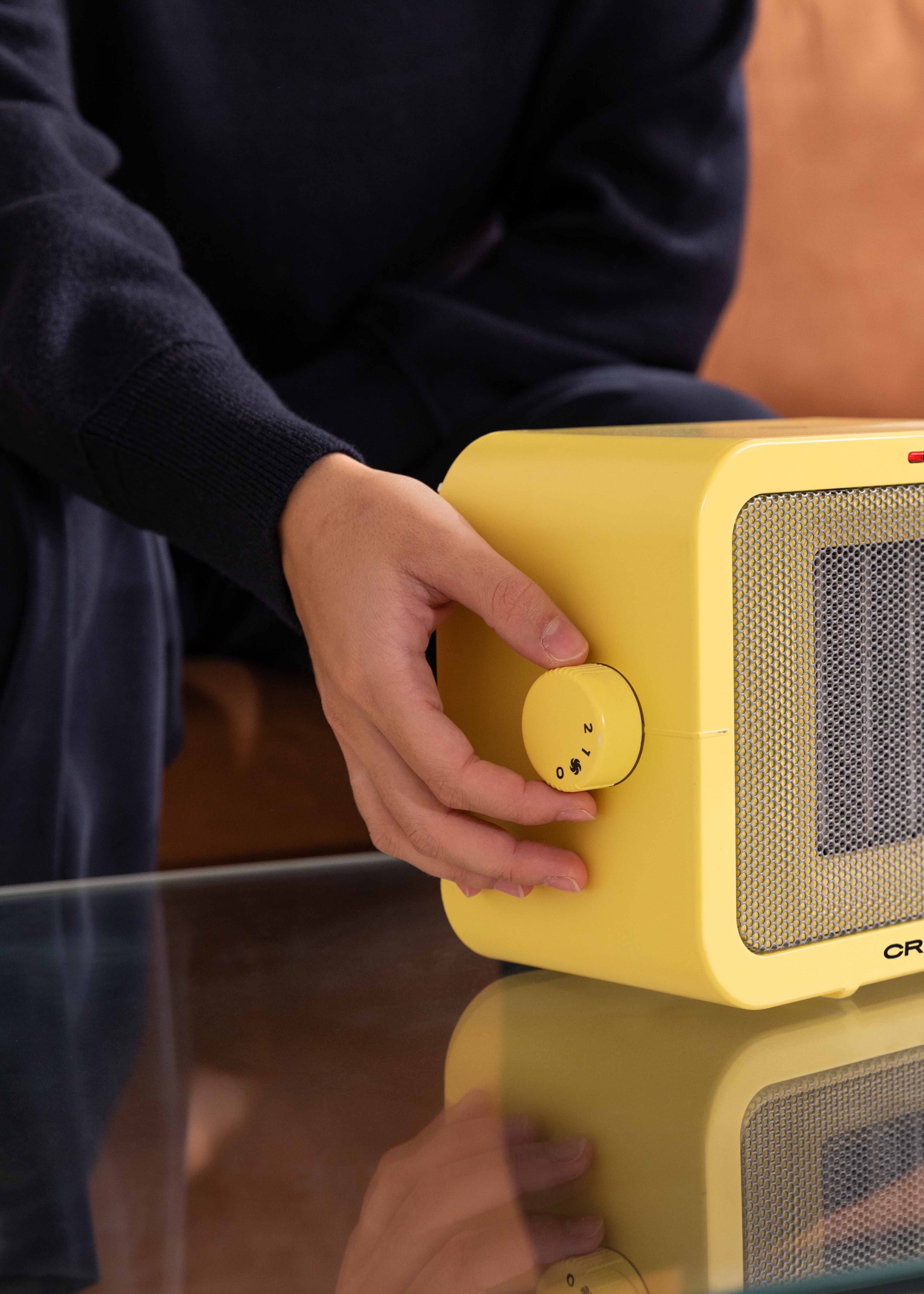 Una persona gira el dial de un calefactor eléctrico amarillo. Mesa de vidrio. Fondo oscuro.