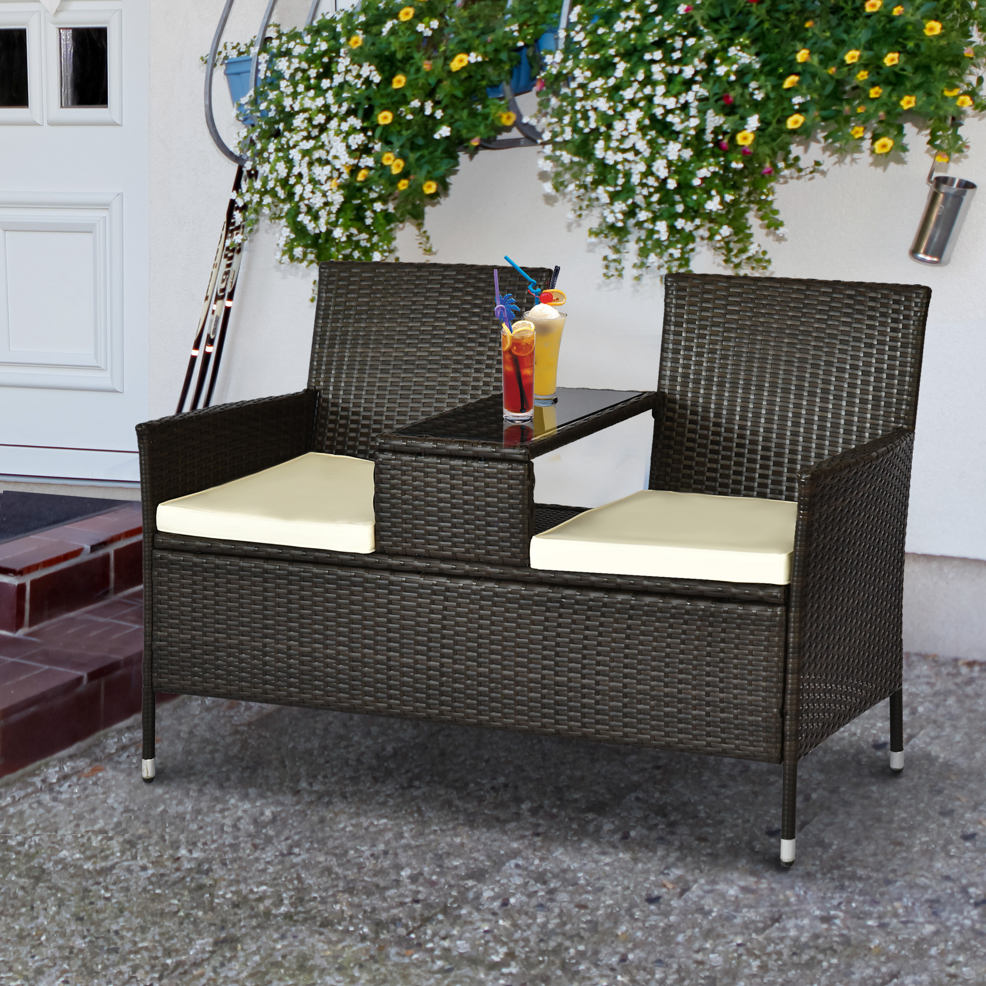 Eine braune Rattanbank mit einem Tisch in der Mitte. Zwei Getränke und Blumen sind zu sehen. Weiße Tür im Hintergrund.