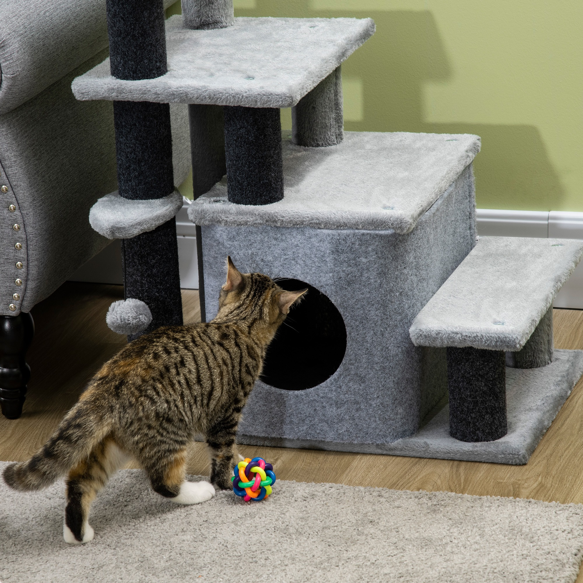 Katze betrachtet Spielzeug auf grauem Katzenbaum. Der Katzenbaum hat mehrere Ebenen und ein Haus.