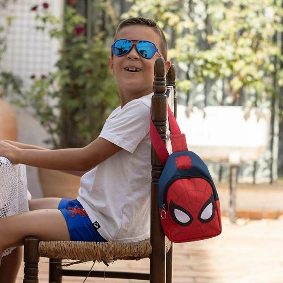 Junger Junge mit Sonnenbrille, trägt eine Spider-Man Umhängetasche. Er lächelt draußen.