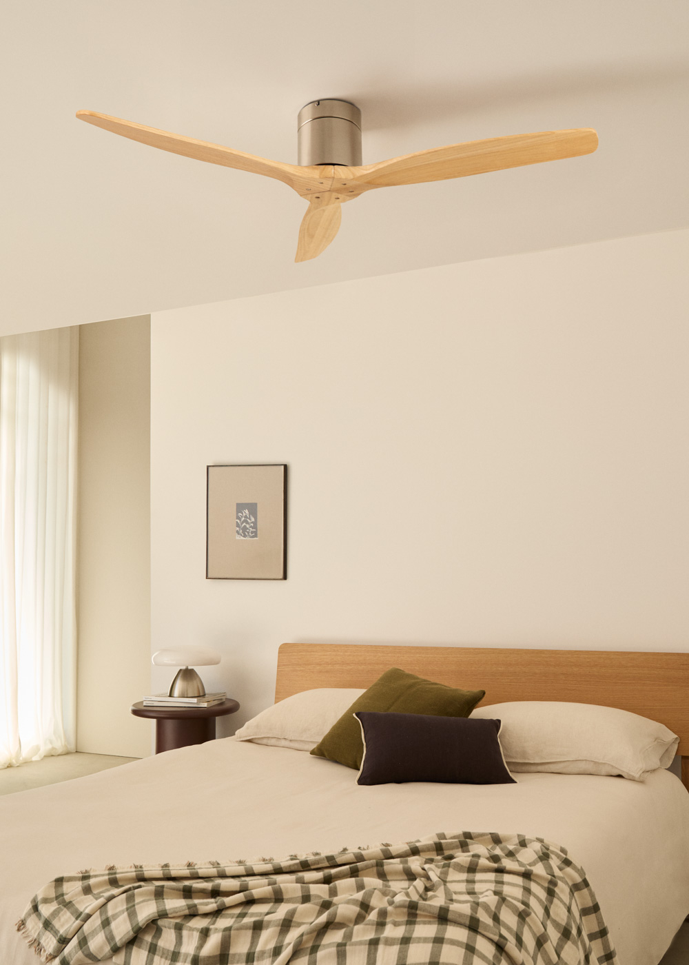 Ventilador de techo de tres aspas de madera, motor plateado, sobre cama. Fondo de pared beige.