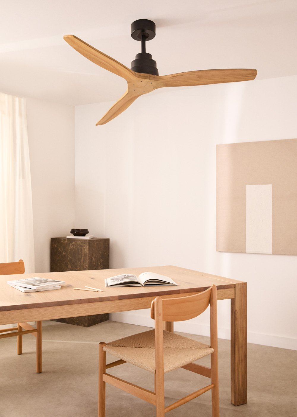 Una mesa de madera con sillas, un ventilador y una pintura en una habitación. El fondo es blanco.