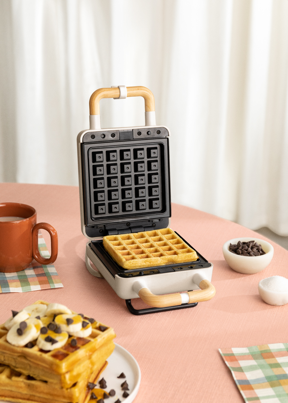 Máquina de hacer gofres con un gofre dentro. Gofres y coberturas en la mesa. Taza marrón.