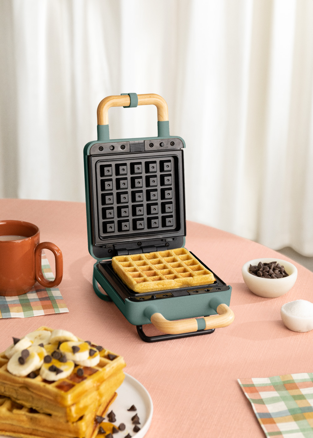 Wafflera verde con un gofre dentro. Plátanos y chispas de chocolate en gofres en la mesa.